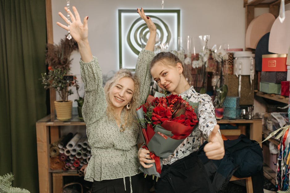 Two young women inside a floral shop holding a vibrant bouquet of red roses wrapped in grey paper along with other red flowers, celebrating with smiles and cheerful expressions. One woman, with blonde
