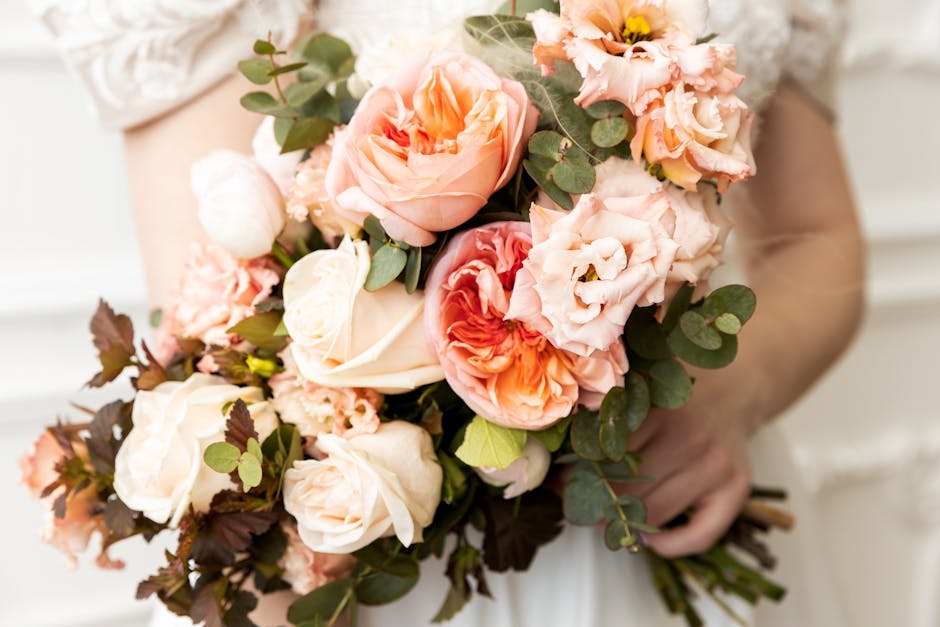 A close-up of a bride holding a lush bouquet of soft peach and cream roses, complemented by delicate peach lisianthus and pale pink peonies. The arrangement features green eucalyptus leaves and dark f