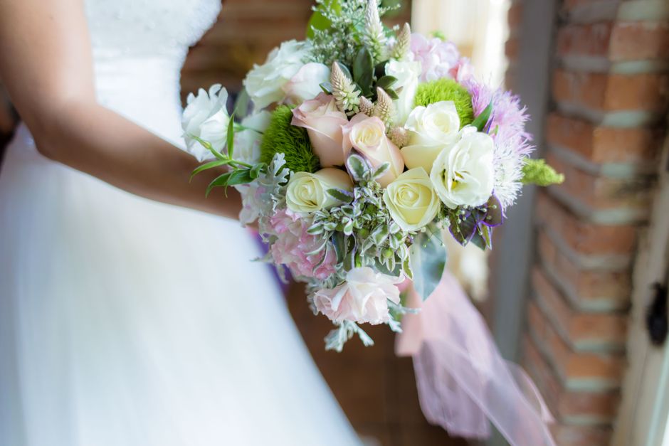 A bridal bouquet featuring cream and blush pink roses, white lisianthus, and pastel pink hydrangeas arranged with green foliage, wrapped in soft pink tulle ribbon. The bouquet is held by a bride weari