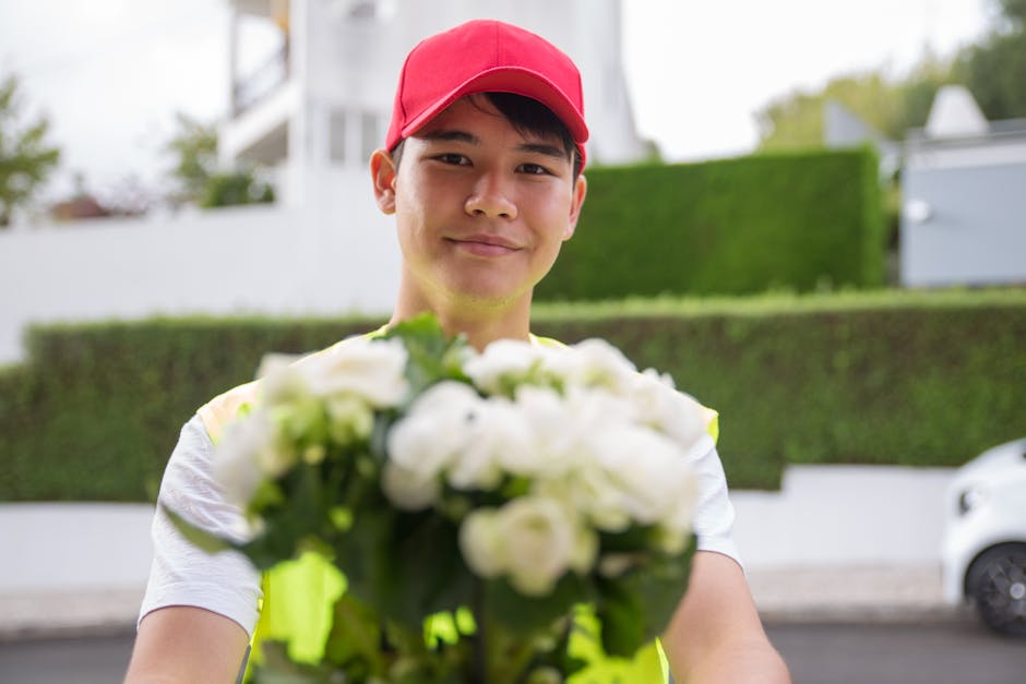 A young delivery person wearing a red cap and a high-visibility vest is holding a bouquet of fresh white roses, which are arranged with green foliage and wrapped in clear plastic. The individual is st