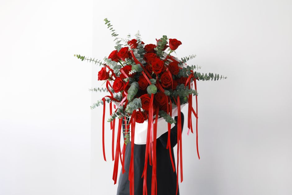 A bouquet arrangement featuring numerous deep red roses with lush, velvety petals, complemented by sprigs of silver-green eucalyptus and other greenery. The flowers are artistically assembled in a rou