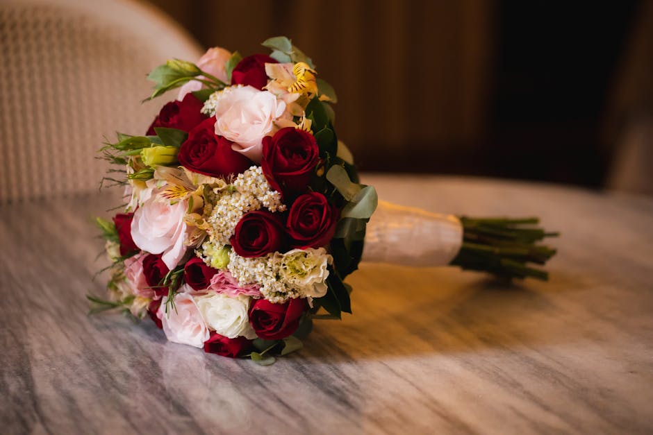 A bouquet arrangement of fresh flowers resting on a wooden surface, featuring deep red roses, soft pink roses, white lisianthus, and delicate white and yellow Alstroemeria, complemented by lush green 