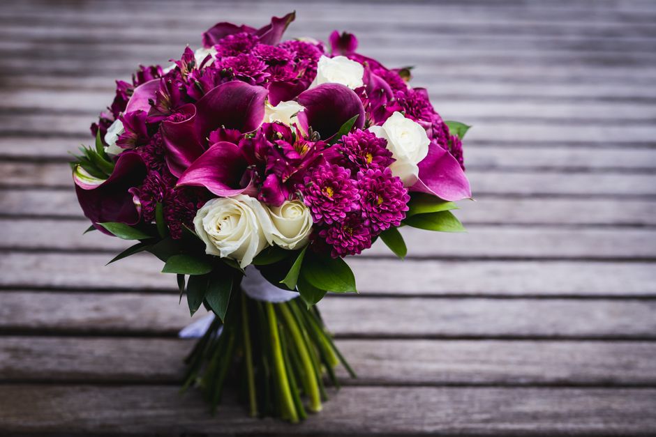 A bouquet of fresh flowers arranged with deep magenta calla lilies, violet chrysanthemums, and white roses, all with vibrant, healthy petals and green foliage, wrapped in white paper and tied with a r