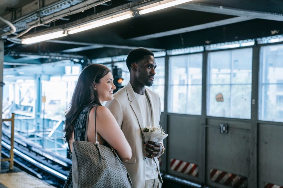 A woman and a man stand on a platform at Southbury station, with the woman smiling and holding a large, colorful bouquet of fresh flowers wrapped in clear plastic and purple paper. The bouquet feature