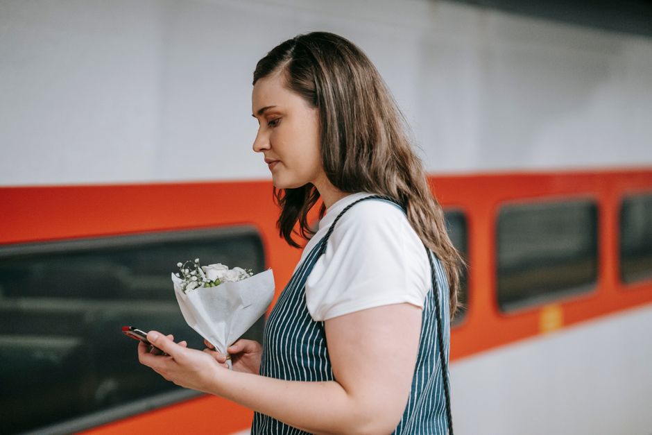A woman with long brown hair, dressed in a white t-shirt and navy and white striped apron, is holding a small bouquet of fresh white flowers wrapped in white paper and tied with a white ribbon. She st